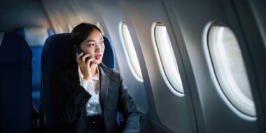 Başarılı Asyalı iş kadını, uçakta dizüstü bilgisayarla çalışan iş kadını ve telefonla konuşurken pencereden dışarı bakan..