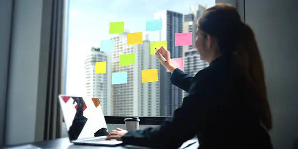 Business woman writing idea or tasks on sticky paper on wall, female team leader, executive manager, planning project, organize work.
