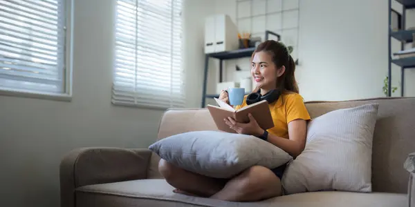 快乐的年轻女人在家里的沙发上看书 生活方式自由职业在客厅里休息 生活放松的概念 — 图库照片