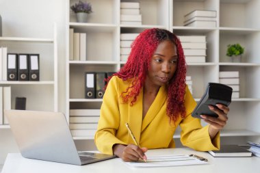 Muhasebeci Afrikalı genç iş kadını muhasebe muhasebesi için hesap makinesi kullanıyor bilgisayarın başında dizüstü bilgisayarın başında oturuyor..