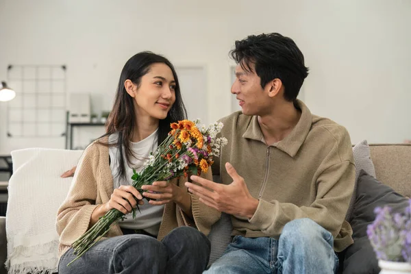 stock image Romantic young asian couple embracing with holding flowers and smiling in living room at home. fall in love. Valentine concept.