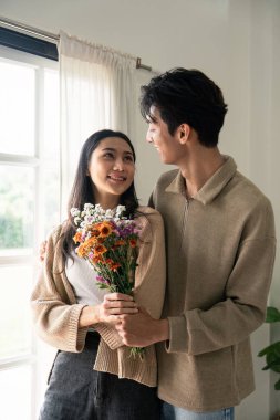 Romantik, genç Asyalı çift ellerinde çiçeklerle oturma odasında gülümsüyor. Aşık olmak. Valentine konsepti.