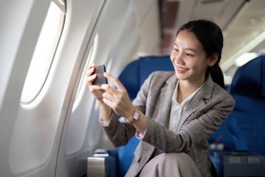Asyalı genç iş kadını başarılı ya da resmi takım elbiseli kadın girişimci birinci sınıf koltukta oturuyor ve uçuş sırasında akıllı telefon kullanıyor. Seyahat ve İş Konsepti.