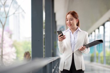 Akıllı telefon ve belge dosyasıyla güzel Asyalı iş kadını, akıllı telefon yürüyüşü yapan Asyalı kadın işe gidiyor. Çalışma günü için hazırlık.