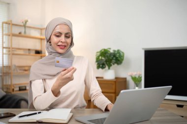 Evde tesettürlü genç mutlu müslüman kadın masada otururken kredi kartıyla online laptop alışverişi yapıyor..