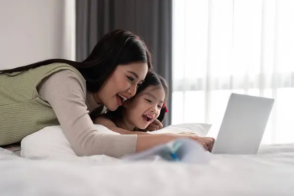 Mother and daughter lying in bed with on and smiling. Young mom working from home in bedroom with laptop.