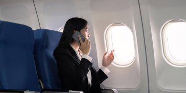 Asyalı genç iş kadını başarılı ya da resmi takım elbiseli kadın girişimci birinci sınıf koltukta oturuyor ve uçuş sırasında akıllı telefon kullanıyor. Seyahat ve İş Konsepti.
