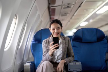 Asyalı genç iş kadını başarılı ya da resmi takım elbiseli kadın girişimci birinci sınıf koltukta oturuyor ve uçuş sırasında akıllı telefon kullanıyor. Seyahat ve İş Konsepti.