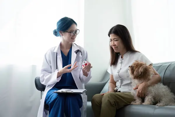 獣医クリニックでアジア女性獣医師の前で机に座っている犬を持つ高齢女性 — ストック写真