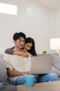 Laptop kullanan mutlu Asyalı çift kanepede oturup evdeki web sitesinde ekommerce alışverişi yapıyor..