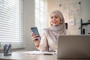 İşadamı Müslüman bayan girişimci ofiste dizüstü bilgisayarla çalışıyor, cep telefonuyla iş görüşmesi yapıyor..