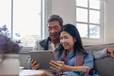 Tablet kullanan mutlu yaşlı Asyalı çift kanepede oturup internet sitesinden ekommerce alışverişi yapıyor ve evde sigorta broşürü satın alıyor..