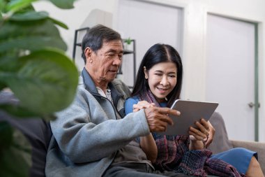 Tablet kullanan mutlu yaşlı Asyalı çift kanepede oturup internet sitesinden ekommerce alışverişi yapıyor ve evde sigorta broşürü satın alıyor..