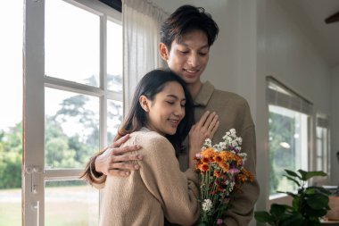 Romantik, genç Asyalı çift ellerinde çiçeklerle oturma odasında gülümsüyor. Aşık olmak. Valentine konsepti.