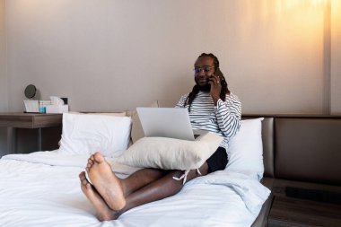 Genç Afro-Amerikalı adam cep telefonuyla konuşuyor ve evde çalışırken laptop ekranına bakıyor..