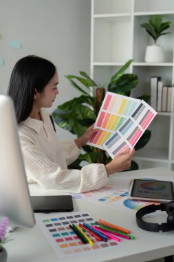 Asyalı kadın serbest grafik tasarımcısı ev ofisindeki masada renk örnekleri ve bilgisayarla çalışıyor, genç bayan yeni tasarım projesi için renk gaması seçiyor.