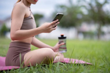 Atletizmde genç bir kadın parkta yoga yapıyor, akıllı telefon kullanıyor, su şişesi tutuyor, sağlıklı bir yaşam tarzı ve kondisyonu destekliyor..