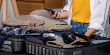 Giysi ve aletlerle dolu bir bavul hazırlayan, online uçuş ve otel rezervasyonu için akıllı telefon kullanan biri. Yaz tatili ve seyahat hazırlığı düşüncesi.