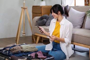 Yaz tatili için uçak bileti ve otel ayırtmak için kredi kartı ve akıllı telefon kullanan genç bir kadın. Konforlu oturma odasında bavulla yerde oturmak, internetten gezi planlamak..