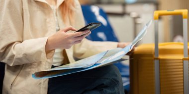 Havaalanında bavulu ve haritası olan genç bir kadın akıllı telefonunu seyahat planlama ve navigasyon için kullanıyor. Modern seyahat temaları, yalnız kadın gezginler ve seyahat hazırlıkları için ideal..