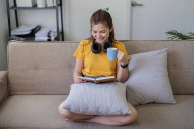 Kanepede kitap okuyan genç bir kadın, bir fincan kahve içip keyfine bakıyor, günlük giysiler ve kulaklık takıyor..