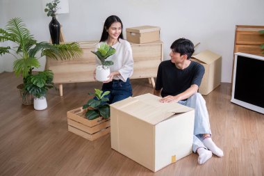 Genç bir çift yeni evlerine taşınıyor, kutuları açıyor ve modern bir oturma odasında bitki düzenliyor. Heyecanlı ve mutlular, yeni başlangıçları ve ev sahipliğini simgeliyorlar..