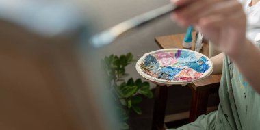 Bir atölyede resim yapan, elinde renkli bir palet ve fırça tutan, stüdyo ortamındaki yaratıcı süreci yakalayan bir sanatçının yakın çekimi..