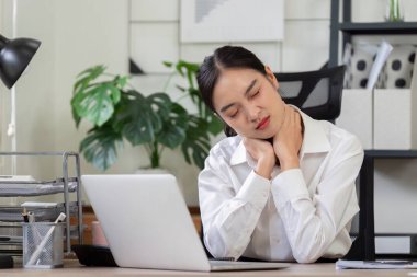 Modern bir ofis ortamında masasında çalışırken boynu ağrıyan beyaz gömlekli bir iş kadını..