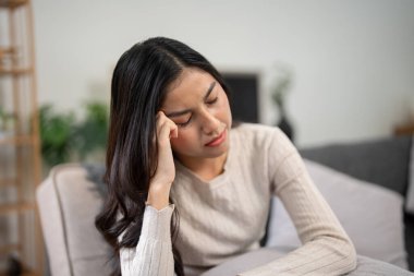 Kanepede oturan genç bir kadın, acı içinde başını tutuyor, baş ağrısı ve migren, hasta ve iyi hissetmiyor..