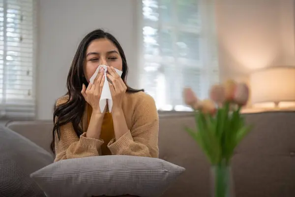 stock image Young woman sitting on a couch sneezing into a tissue due to pollen allergy, with a vase of flowers in the foreground