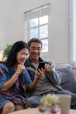 Mutlu yaşlı çift birlikte akıllı telefon kullanıyor, alışveriş ve sosyal medya uygulamalarını keşfediyor evde rahat bir kanepede otururken.