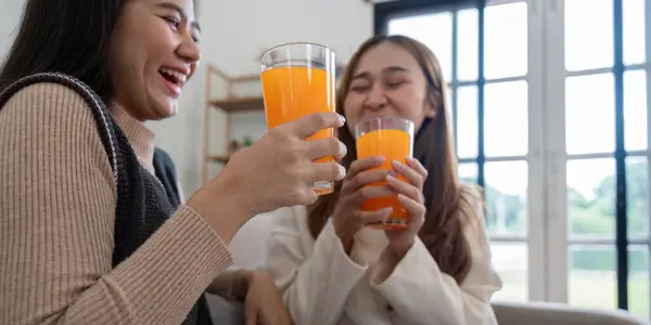 stock image Two smiling women share a moment of joy while drinking healthy orange juice in a cozy, sunlit home environment.