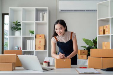 Genç bir iş kadını, çevrimiçi ekommerce ve ürün nakliyesi sürecini gözler önüne sererek, modern bir ev ofisinde teslimat için paket kutuları hazırlıyor..