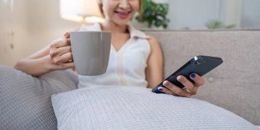 Bir kadın kanepede rahatça oturur, elinde bir kahve fincanı tutar ve sosyal medyada rahat bir şekilde gezinmek için akıllı telefonunu kullanır..