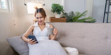 Genç bir kadın rahat bir kanepede rahatlar, kahvesini yudumlar ve sosyal medyada akıllı telefonuyla rahat, modern bir oturma odasında gezinir..