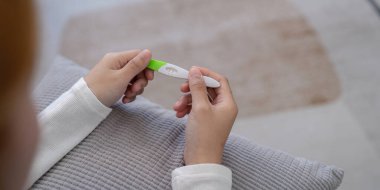 Hamilelik testi kiti tutan ellerin yakın plan fotoğrafı. Pozitif sonuç veriyor, rahat bir ev ortamında ayarlanmış..