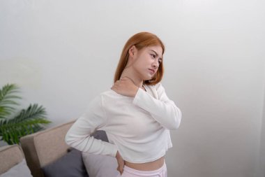 Beyaz bluzlu genç bir kadın, boyun ve sırt ağrısı belirtileri gösteriyor, modern bir ev ortamında duruyor..