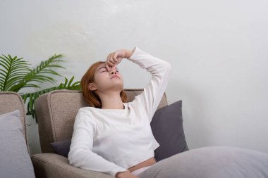 Kanepede oturan, köprüsünü tutan, modern oturma odasında baş ağrısı çeken genç bir kadın..