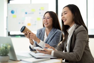 İki profesyonel kadın iş kıyafetleri içinde parlak, modern bir ofiste çalışıyor, fikirleri paylaşıyor ve gülümsüyor..