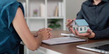 Emlakçı ve müşteri emlak alım-satımı için belge imzalarken, emlakçı elinde bir ev modeli tutuyor..