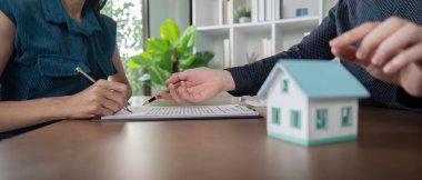 Bir emlakçı, emlak alım-satım sürecini sembolize ederek müşteriye ev satın alma veya satma belgeleri imzalamasında yardımcı olur..