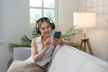 Genç bir kadın kanepede dinlenmekten, kulaklıkla müzik dinlemekten ve rahat, aydınlık bir oturma odasında akıllı telefonunu kullanmaktan hoşlanır..