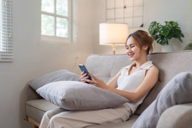 Genç bir kadın, parlak, rahat bir oturma odasında, akıllı telefonuyla sosyal medyayla meşgul olarak kanepede dinleniyor..