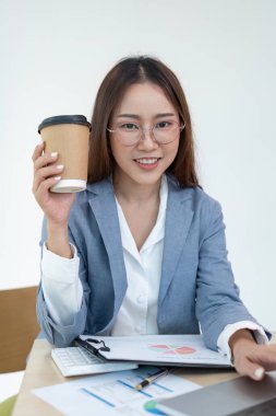 Gözlüklü, elinde kahve fincanı olan, elinde belgelerle bir masada oturan ve modern bir ofiste hesap makinesi olan kendinden emin genç bir iş kadını..