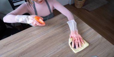 Eldiven takan bir hizmetçi modern bir evde, tahta bir masayı sprey şişeyle ve kumaşla temizliyor..