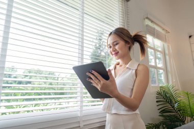 Genç bir kadın evdeki dinlenme anından zevk alıyor. Parlak, modern bir oturma odasında, pencerenin kenarında bir tablet kullanıyor..