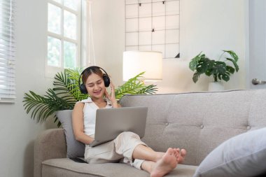 Bir kadın, koltukta oturup, kulaklıkla müzik dinlemekten ve parlak, rahat bir oturma odasında dizüstü bilgisayar kullanmaktan zevk alır..