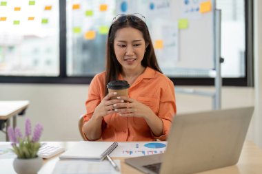 Modern ofis ortamında kahve ve belgelerle dolu bir dizüstü bilgisayarla çalışan, renkli yapışkan notlarla çevrili genç bir kadın..