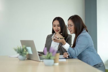 Modern bir ofiste dizüstü bilgisayarda birlikte çalışan iki profesyonel iş kadını bir proje üzerinde tartışıyor ve işbirliği yapıyorlar..