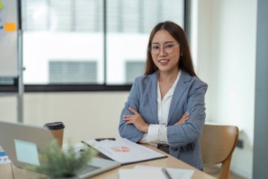 Modern ofis ortamında kolları bağlı, dizüstü bilgisayarı ve evraklarıyla güvenle gülümseyen profesyonel bir iş kadını..